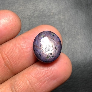 May include: A round, polished gemstone with a deep purple hue, speckled with lighter tones and reflective inclusions. The gem is held between fingers, showcasing its smooth, rounded shape and natural beauty. The stone appears to be a cabochon cut.