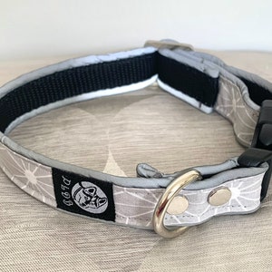 May include: A gray dog collar with a white geometric pattern and a black buckle. The collar has a reflective strip and a silver metal D-ring. A black tag with a dog logo is sewn on the collar.