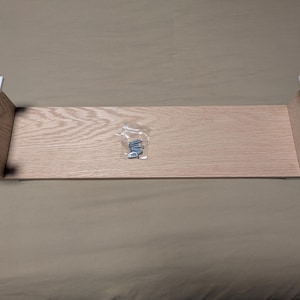 May include: A light-colored wooden shelf with white corner brackets and a small bag of screws. The shelf is rectangular and appears to be new and unassembled. The shelf is made of wood and is ready to be installed.
