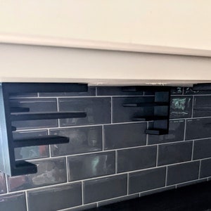 May include: Two black metal shelf units mounted under a white cabinet. The shelves are staggered and create a modern, geometric design. The shelves are mounted on a gray tiled wall.