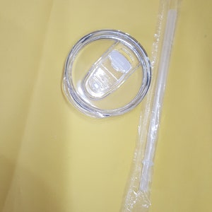 May include: A clear plastic lid with a sliding closure for a tumbler or cup. The lid is on a yellow surface next to a clear plastic straw in a sealed plastic bag.