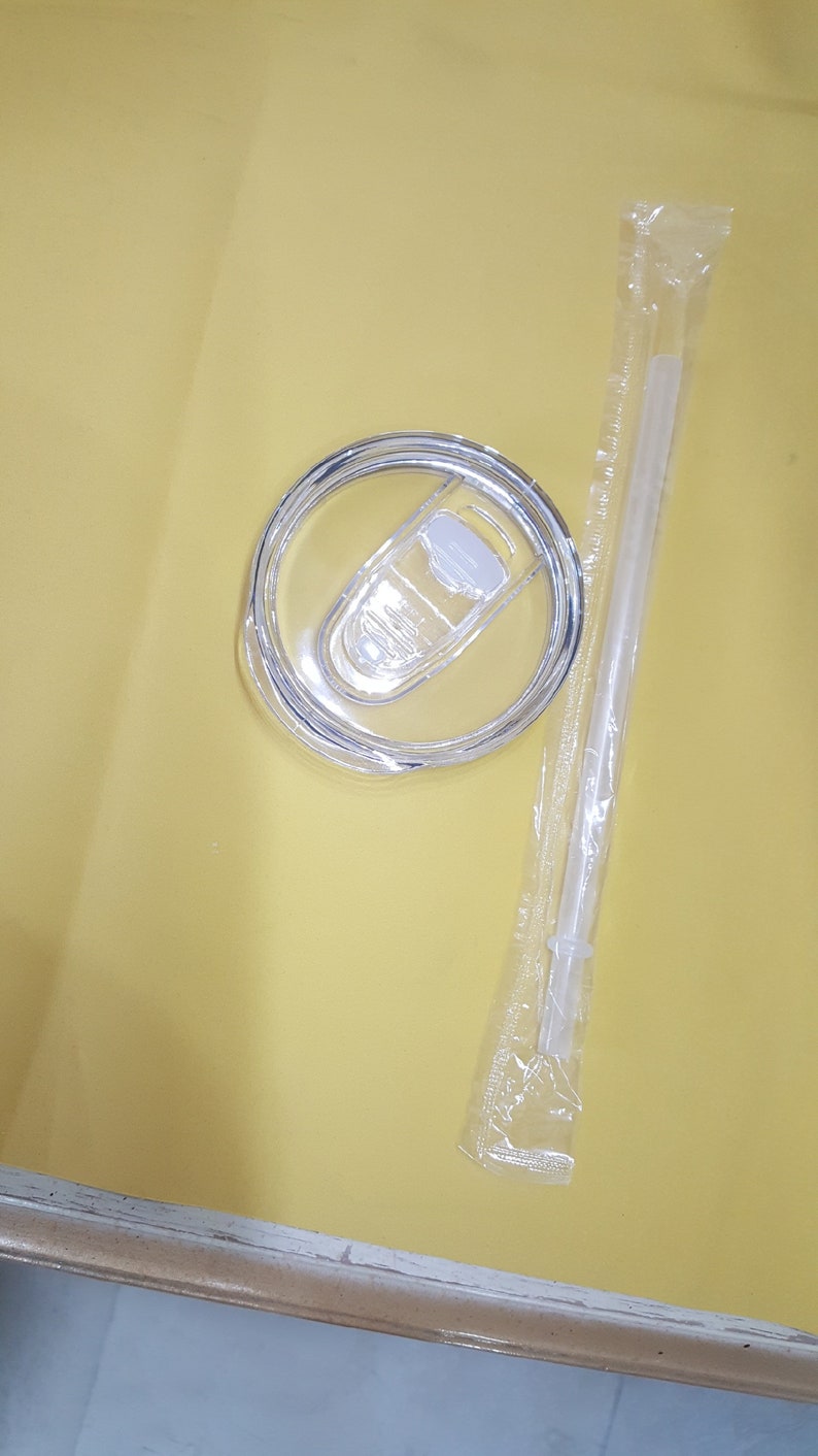 May include: A clear plastic lid with a sliding closure for a tumbler or cup. The lid is on a yellow surface next to a clear plastic straw in a sealed plastic bag.