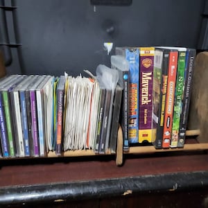 May include: A wooden shelf filled with CDs, DVDs, and VHS tapes. The media includes music CDs, movies like "Maverick" and "Peter Pan", and other titles. The shelf is made of light-colored wood and sits on a dark surface.