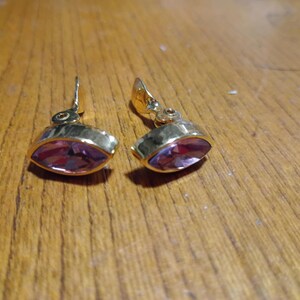 May include: A pair of gold-toned clip-on earrings with a marquise-shaped, purple gemstone center. The earrings are set on a wooden surface, showcasing their design and color.