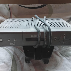 May include: A silver Samsung DirecTV receiver with a perforated top and a black base. The front panel has buttons labeled "POWER", "INFO", and "SAT", along with a directional pad. Cables are coiled on top.