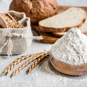 May include: A close-up of a bowl of white flour, a hessian sack filled with wheat grains, and a loaf of bread. The flour is piled high in the bowl, and the wheat grains are spilling out of the sack. The bread is sliced and arranged on a wooden chopping board.