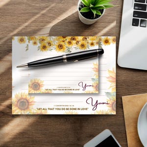 May include: A set of two lined notepads with a sunflower design. The notepads have a white background with a border of sunflowers and green leaves. The top notepad has the text "Let all that you do be done in love" and the bottom notepad has the text "Yumi".