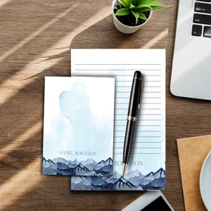 May include: Two personalized stationery sets with a watercolor mountain design. The top sheet features a light blue watercolor wash with a mountain range at the bottom. The bottom sheet features a dark blue mountain range with a light blue watercolor wash at the top. The name "DANIEL BORGESON" is printed on the top sheet. The name "BORGESON" is printed on the bottom sheet.