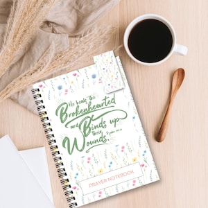May include: A white prayer notebook with a floral pattern and the text "He heals the brokenhearted and binds up their wounds. Psalm 147:3." The notebook has a green spiral binding and says "PRAYER NOTEBOOK" at the bottom.