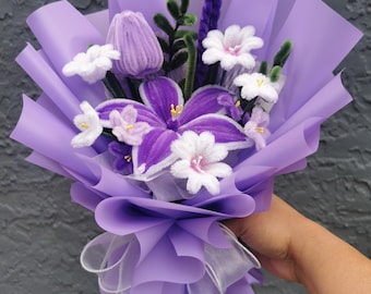 May include: A bouquet of handmade flowers wrapped in lavender paper and tied with a white ribbon. The bouquet features purple and white flowers, including a large violet-like bloom, and a lavender-colored tulip-shaped flower.