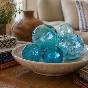 May include: A collection of decorative glass spheres in varying shades of blue, resting in a textured, beige bowl. The spheres have different surface textures, including smooth and speckled. A stack of books with the word "MEXICO" is visible.