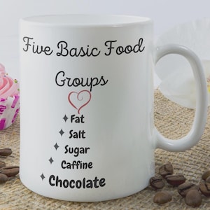 Puede incluir: Taza de cerámica blanca con un diseño en negro y rojo. La taza tiene un asa y dice "Five Basic Food Groups" con un corazón y una lista de elementos: Fat, Salt, Sugar, Caffine, Chocolate.