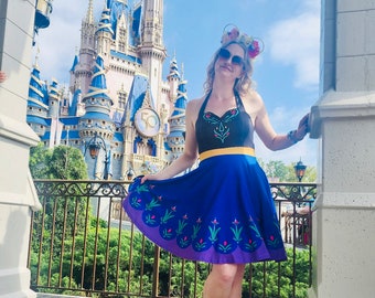 May include: A woman wearing a blue and black dress with a floral design, yellow belt, and floral Minnie Mouse ears stands in front of Cinderella's Castle. The dress has a blue skirt with a purple hem and green and pink floral accents.