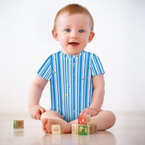 May include: A baby wearing a blue and white striped bodysuit sits on the floor, smiling. The bodysuit has a pattern of vertical stripes and small colorful shapes. Wooden alphabet blocks are scattered in front of the baby.