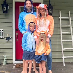 May include: A family of four wearing matching animal-themed hoodies. The father is wearing a blue hoodie with a white and orange design, the mother is wearing an orange hoodie with a white and brown design, and the two children are wearing blue and orange hoodies with animal ears. They are standing on a wooden porch in front of a red door.