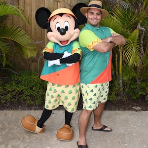 May include: Mickey Mouse wearing a green and orange shirt and shorts with a yellow and green pattern. He is standing next to a person wearing a green and yellow shirt and shorts with a yellow and green pattern. Both are wearing hats.