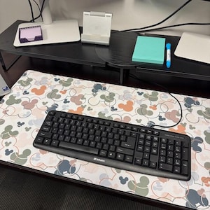 May include: A rectangular desk mat featuring a repeating pattern of overlapping circles and mouse-ear silhouettes in muted shades of sage green, dusty rose, and slate blue on a white background, with a black wired computer keyboard placed on top.