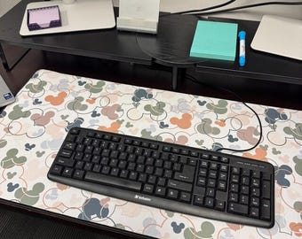 May include: A rectangular desk mat featuring a repeating pattern of overlapping circles and mouse-ear silhouettes in muted shades of sage green, dusty rose, and slate blue on a white background, with a black wired computer keyboard placed on top.
