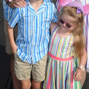 May include: Two children wearing striped clothing. The boy is wearing a blue and white striped shirt with a beige pair of shorts. The girl is wearing a multi-colored striped dress with a pink sash and a pink and purple headband with a crown.
