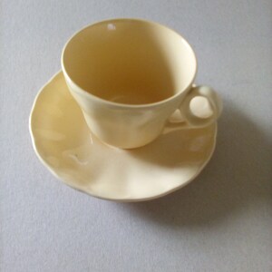 May include: A pale yellow teacup and saucer set. The cup has a rounded shape and a small handle. The saucer has a scalloped edge.