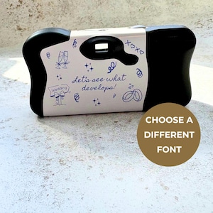 May include: A black disposable camera with a white wrap featuring blue illustrations of champagne flutes, wedding rings, and the text Let's see what develops! The camera has a shutter button on top and a viewfinder window on the front side.