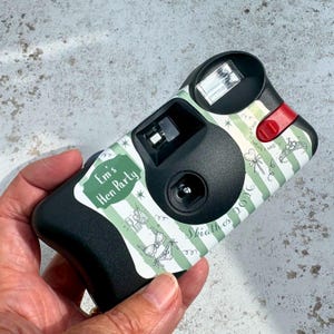 May include: A black disposable camera with a custom green and white striped wrap featuring the text Em's Hen Party and Skiathos 2026. The design includes illustrations of a tiara, a lollipop, and lingerie, with a red flash button on the top right.