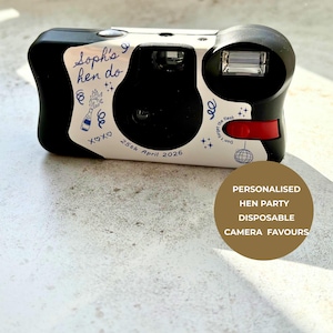 May include: A black disposable camera with a white custom wrap featuring blue line art of a champagne bottle, a disco ball, and stars. The text reads "Soph's hen do", "25th April 2026", and "Don't forget the flash". It includes a red shutter button.