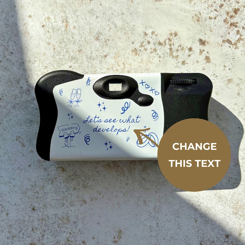 May include: A disposable camera with a white front panel featuring blue line art of champagne flutes, wedding rings, and the text Let's see what develops! A brown circle with the text CHANGE THIS TEXT overlays the right side of the camera.