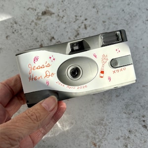 May include: A silver disposable camera with a white custom sticker featuring red text that reads Jess's Hen Do and 25th April 2026. The sticker includes red line art of a champagne bottle, confetti, stars, and the text XOXO on the front.