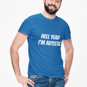 Op de afbeelding: Een blauw t-shirt met de tekst "HELL YEAH I'M AUTISTIC" in het wit. Het shirt wordt gedragen door een persoon in een spijkerbroek. De persoon lacht en heeft een baard.