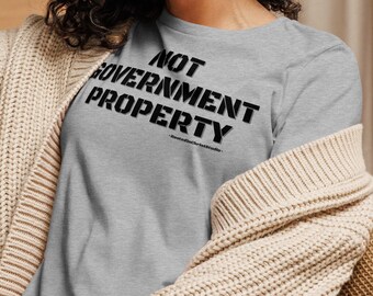 May include: A woman wearing a heather grey t-shirt with the text "NOT GOVERNMENT PROPERTY" printed in black. She is smiling and wearing a beige cardigan.
