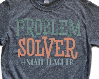 Peut inclure: T-shirt gris foncé avec l'inscription "PROBLEM SOLVER" en vert et orange, et le hashtag "#MATHTEACHER". Le t-shirt est de style décontracté, avec des manches courtes et un col rond.