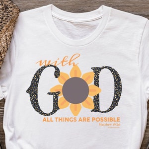 Puede incluir: Camiseta blanca con la frase "with GOD" y un diseño de girasol. El texto "ALL THINGS ARE POSSIBLE Matthew 19:26" está impreso debajo. La camiseta se combina con pantalones cortos de mezclilla, gafas de sol y un sombrero.