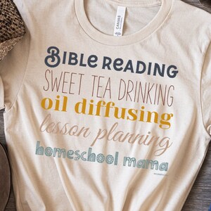 May include: A cream-colored t-shirt with the words "Bible Reading, Sweet Tea Drinking, Oil Diffusing, Lesson Planning, Homeschool Mama" in various colors. A brown cowboy hat, brown suede boots, and blue jeans are also in the image.