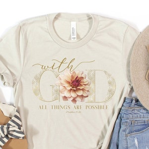 Puede incluir: Una camiseta beige con un diseño floral y el texto "with God all things are possible" en letras doradas. La camiseta se combina con pantalones cortos de mezclilla, un sombrero de paja y sandalias.