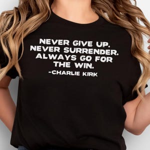 May include: Black t-shirt with the text "NEVER GIVE UP. NEVER SURRENDER. ALWAYS GO FOR THE WIN. -CHARLIE KIRK" in white. The shirt is paired with blue jeans.