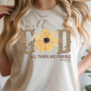 Puede incluir: Una camiseta blanca con un diseño de estampado de leopardo que dice "con Dios todo es posible" y un gráfico de girasol. El texto "Mateo 19:26" también está incluido en la camiseta.