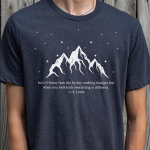 May include: A dark heather grey t-shirt featuring a white mountain range graphic and a quote by C.S. Lewis: "Isn't it funny how day by day nothing changes but when you look back everything is different."