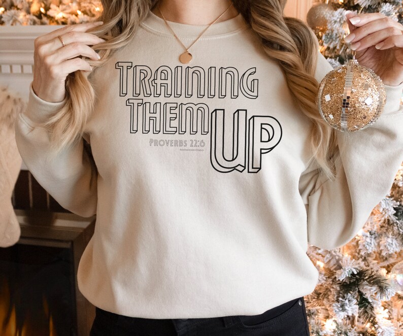 以下が含まれることがあります： クリーム色のスウェットシャツに、黒い輪郭の文字で「TRAINING THEM UP」と書いてあります。「PROVERBS 22:6」という文字も黒で書かれています。