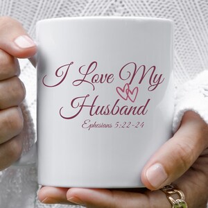 May include: White ceramic mug with the text "I Love My Husband" in burgundy script with two pink hearts. The text "Ephesians 5:22-24" is also in burgundy script.