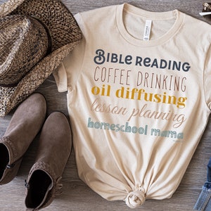 May include: A beige t-shirt with the text "Bible Reading Coffee Drinking Oil Diffusing Lesson Planning Homeschool Mama" printed in various colours. The shirt is tied in a knot at the bottom.