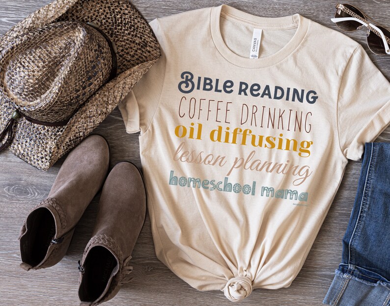 May include: A beige t-shirt with the text "Bible Reading Coffee Drinking Oil Diffusing Lesson Planning Homeschool Mama" printed in various colours. The shirt is tied in a knot at the bottom.