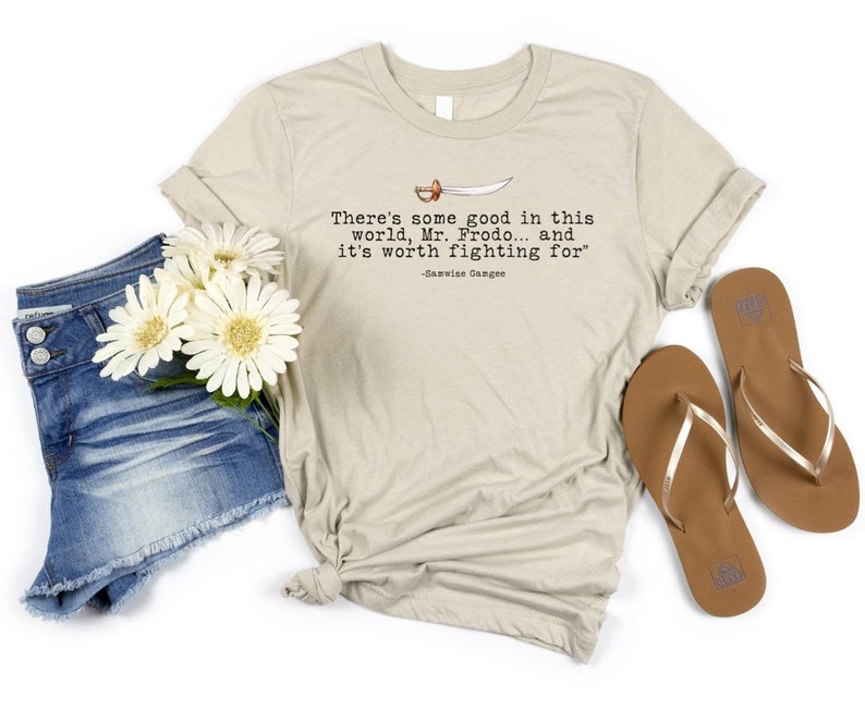 May include: A beige t-shirt with the quote 'There's some good in this world, Mr. Frodo... and it's worth fighting for' -Samwise Gamgee printed on it. The t-shirt is laid out on a white surface with a pair of brown flip flops and a pair of blue denim shorts with white flowers.