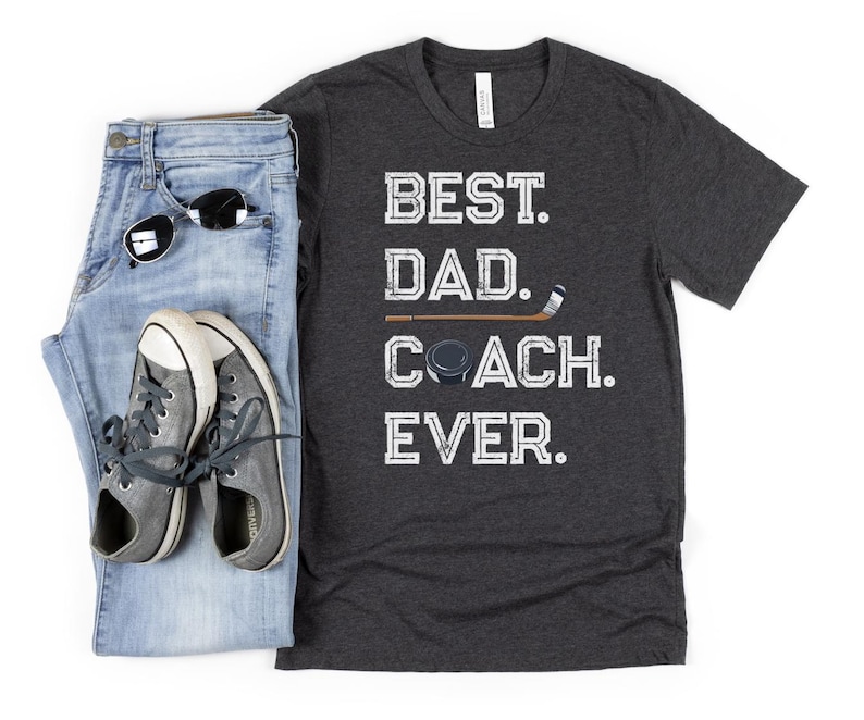 May include: A dark gray t-shirt with the text 'BEST. DAD. COACH. EVER.' in white. The text is in a distressed font. A hockey stick and puck are above the text. The t-shirt is displayed on a white background with a pair of blue jeans and a pair of white sneakers.