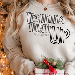 以下が含まれることがあります： 黒い輪郭の文字で「Training Them Up」と書かれたベージュのスウェットシャツ。黒い文字で「箴言 22:6」も書かれています。
