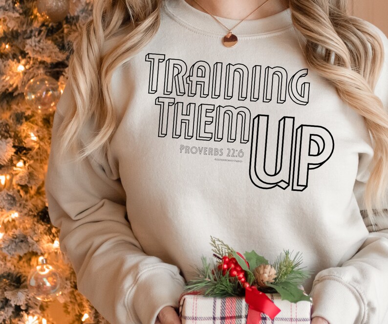 以下が含まれることがあります： 黒い輪郭の文字で「Training Them Up」と書かれたベージュのスウェットシャツ。黒い文字で「箴言 22:6」も書かれています。