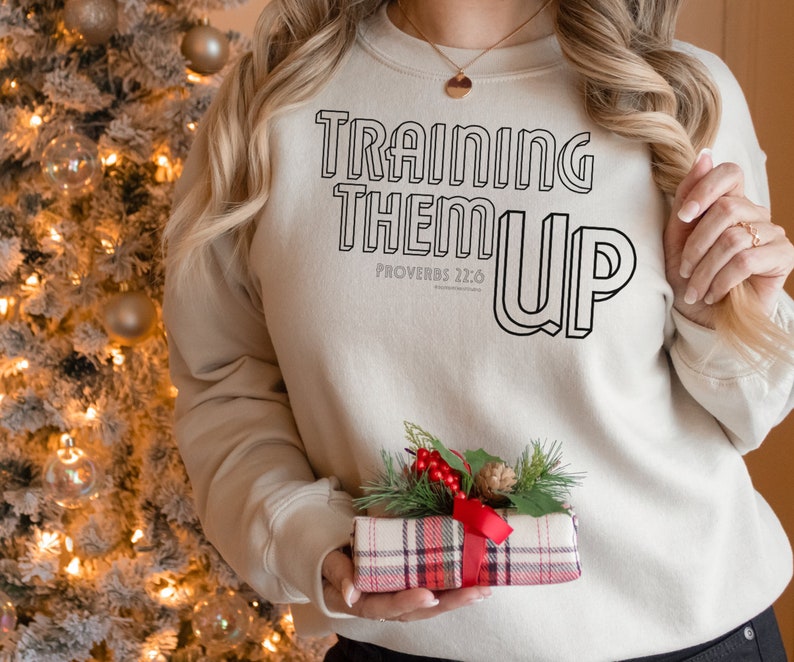 以下が含まれることがあります： 黒い輪郭の文字で「TRAINING THEM UP」と書かれたベージュのトレーナー。黒い文字で「PROVERBS 22:6」も書かれている。