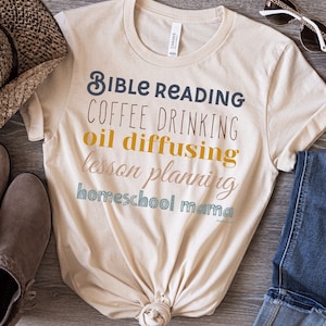 Puede incluir: Una camiseta beige con el texto "Bible Reading, Coffee Drinking, Oil Diffusing, Lesson Planning, Homeschool Mama" en varios colores.