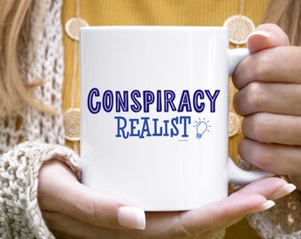 May include: White ceramic mug with a blue outline of the text "Conspiracy Realist" and a lightbulb graphic.