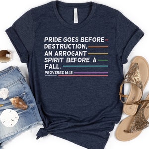 May include: Navy blue t-shirt with the text "PRIDE GOES BEFORE DESTRUCTION, AN ARROGANT SPIRIT BEFORE A FALL. PROVERBS 16:18" in white. The text is underlined with colorful horizontal lines. The shirt is knotted at the waist.